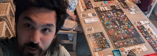 Mickey is smiling on the left of a selfie, with the tabletop game Arcs in the background.