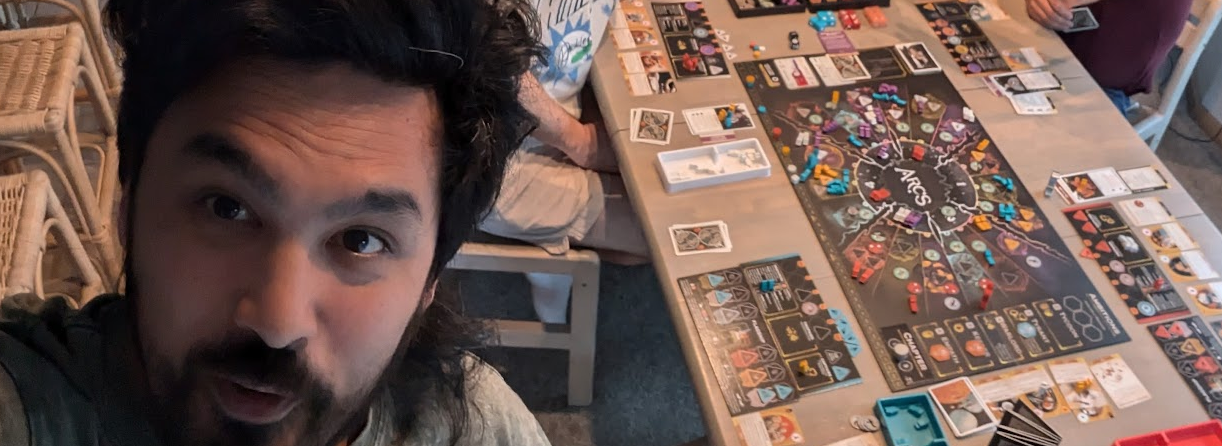 Mickey is smiling on the left of a selfie, with the tabletop game Arcs in the background.